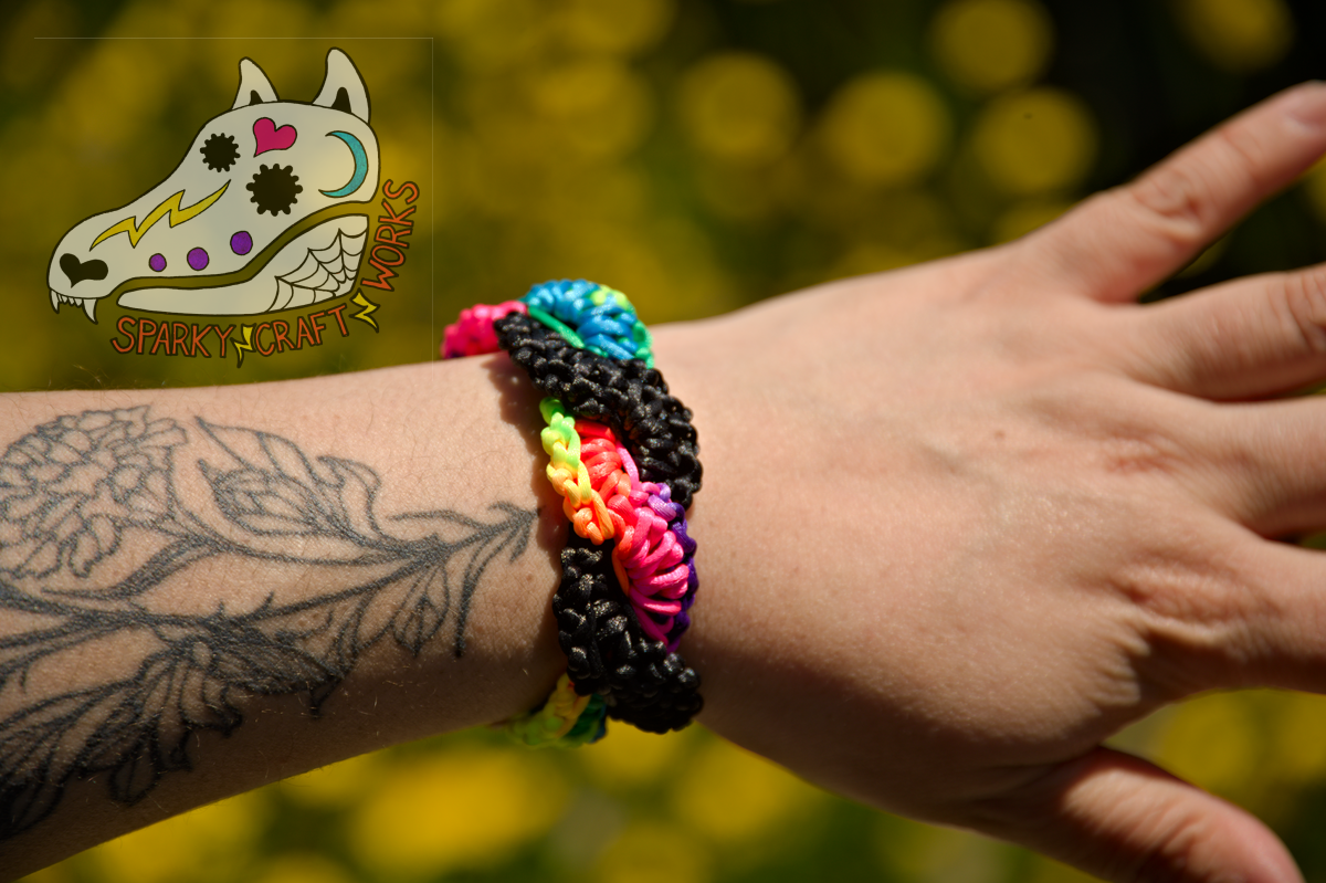 A photo of a rainbow bracelet and black bracelet intertwined, being worn by a brown person with a marigold tattoo going up from their wrist. In the background is blurry green-yellow plants. On the upper-right is the Sparky Craft Works watermark, a stylized calavera-painted wolf skull design.