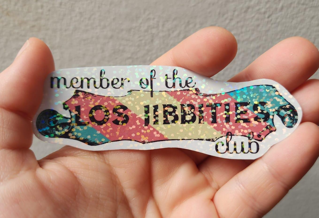 A holographic sparkly rainbow sticker that reads 'member of the LOS JIBBITIES club' on a trans pride flag coloured scroll