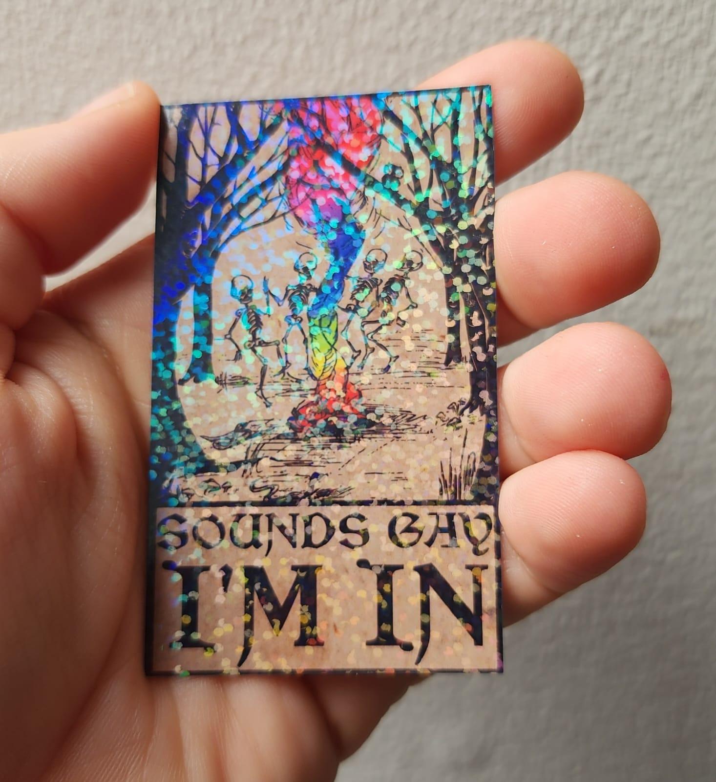 My hand holding up a rectangular rainbow glitter holographic sticker. Four skeletons dance around a rainbow fire in a forest as some woodland critters look on. Text beneath reads 'Sounds gay, I'm in'.