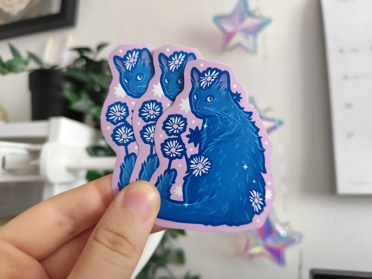 Hand holding up three stickers of a black cat with daisies around it