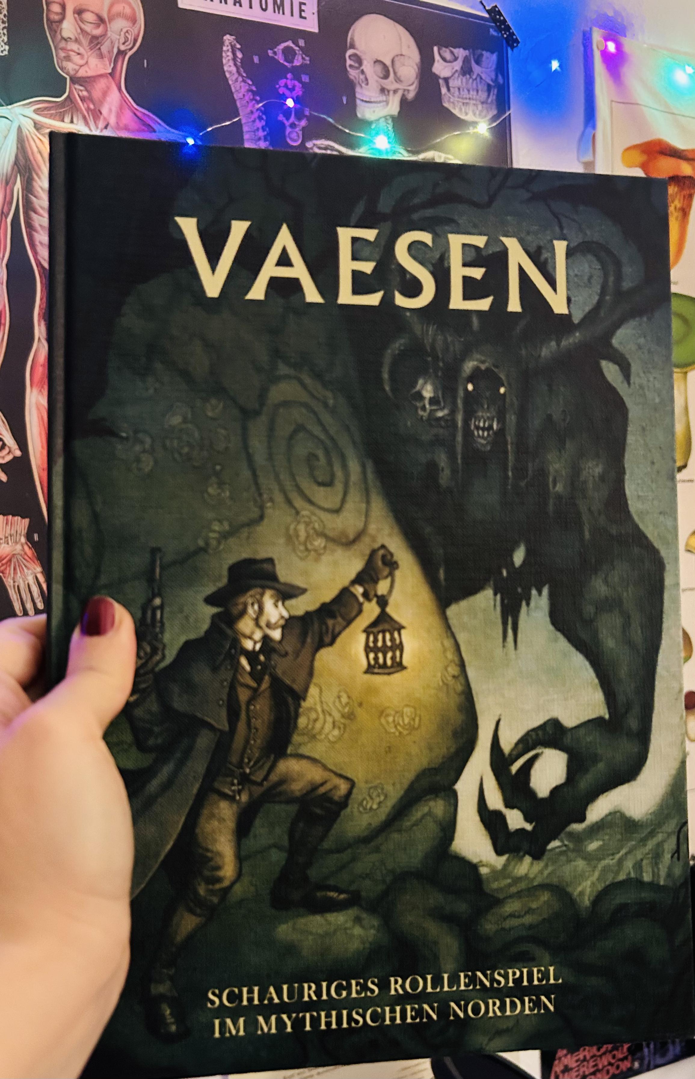 Foto: meine Hand hält das Regelwerk von Vaesen hoch, das Cover ist grün mit einem Mann in viktorianischer Kleidung, der eine Laterne und eine Pistole hält. Daneben steht ein gehörntes Monster.