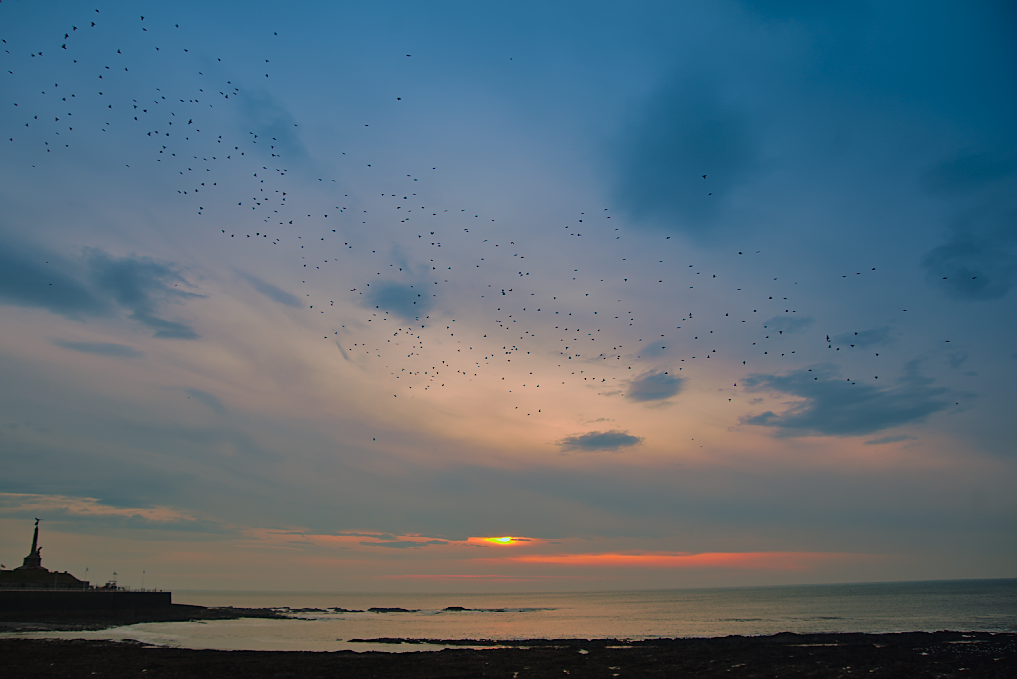 A narrow strip of rocky beach and a pier with a monument in the foreground against a grey-blue, orange-streaked sunset sky. There's a ribbon of starling silhouettes against the clouds, above the water.