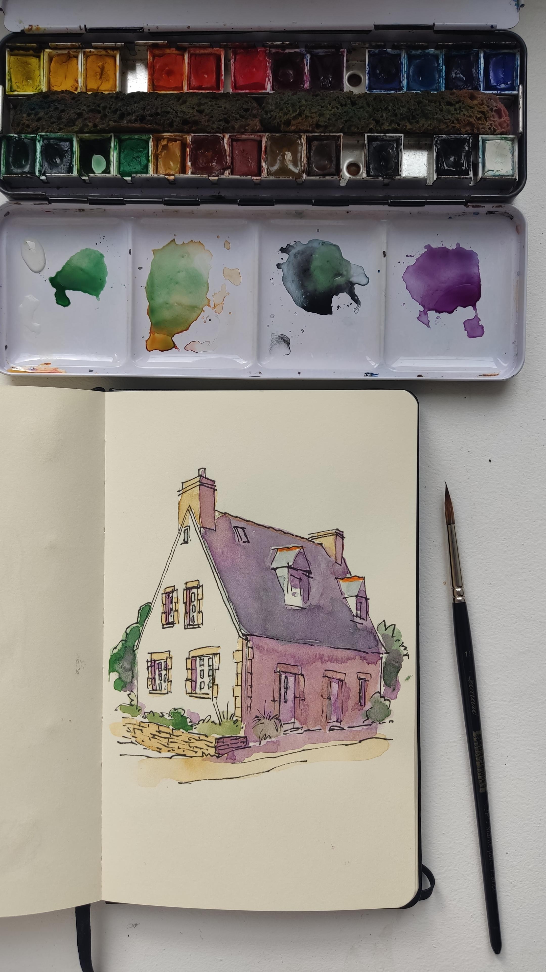 Vue du dessus d'une page de carnet avec, dessiné dessus à l'aquarelle, une maison de style néo breton. Au dessus du carnet, une boîte de couleurs, et à droite un pinceau.