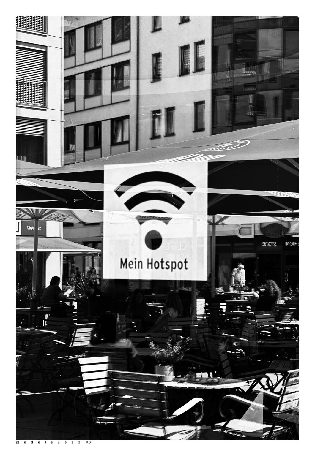 Aufnahme in schwarzweiß, eine große Schaufensterscheibe, darauf das WIFI Symbol und der Schriftzug „Mein Hotspot“, in dem Fenster spiegelt sich eine Außenfläche eines Cafés mit Tischen, Stühlen und Sonnenschirmen, unscharf einige Gäste sitzend, darüber ein eckiges Gebäude mit vielen Fenstern
