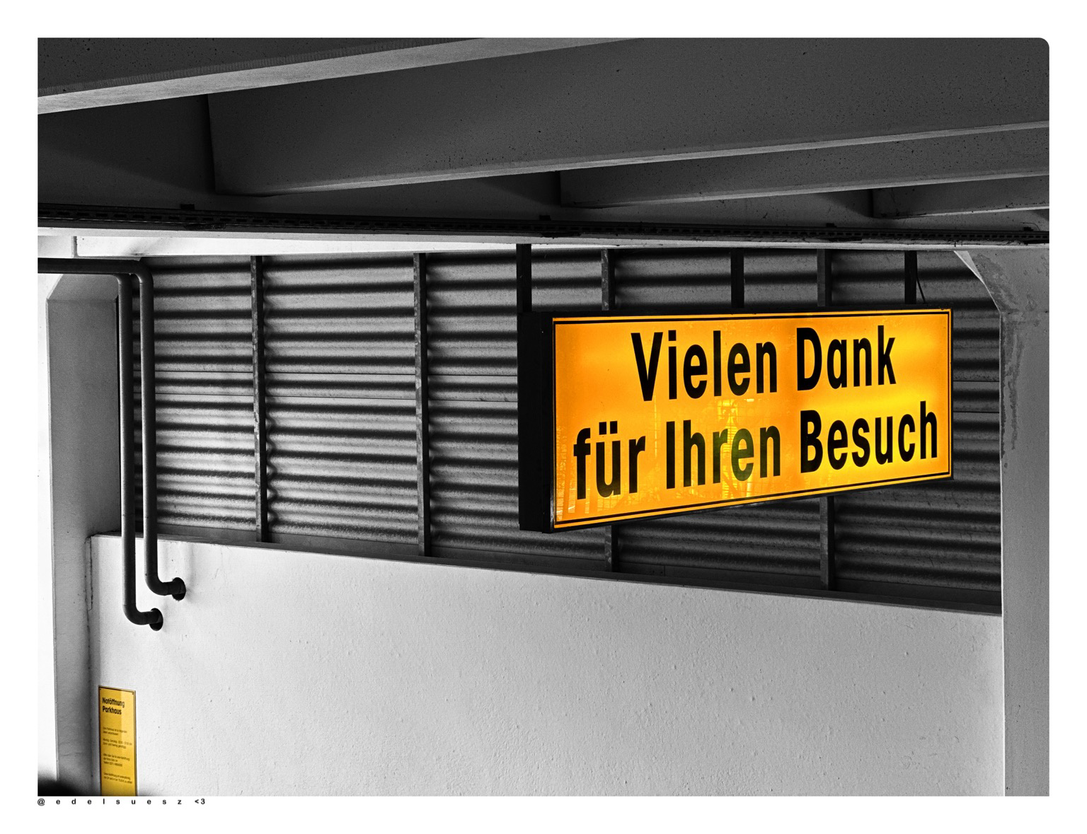 Ausfahrt aus einem Parkhaus, ein beleuchtetes Schild über der Ausfahrt „Vielen Dank für Ihren Besuch“, dieses ist orangegelb, der Rest des Bildes ist schwarzweiß gehalten
