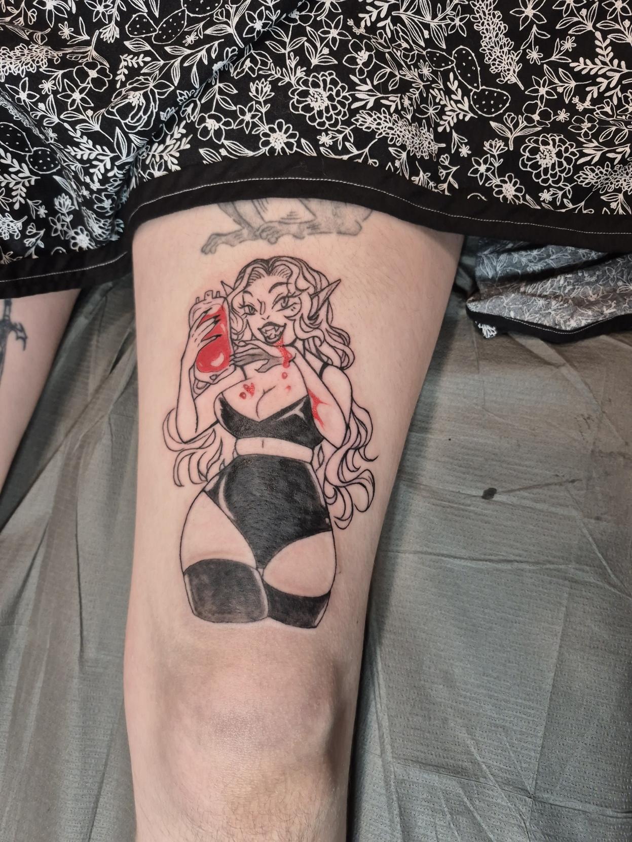 Photo of a freshly done colour pop black and grey tattoo, it's a pin up of a voluptuous vampire lady enjoying a drink from a blood bag. She wears a shiny black bikini top, booty shorts, and stockings.