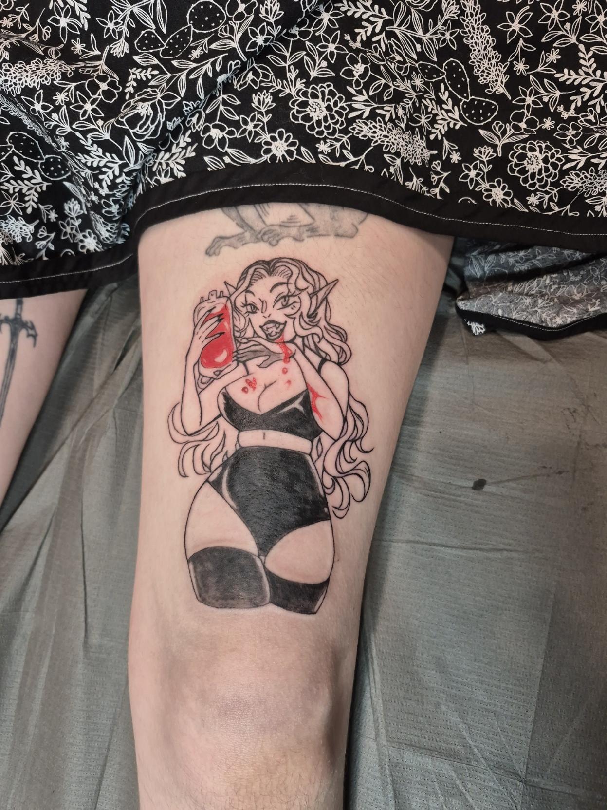 Photo of a freshly done colour pop black and grey tattoo, it's a pin up of a voluptuous vampire lady enjoying a drink from a blood bag. She wears a shiny black bikini top, booty shorts, and stockings.