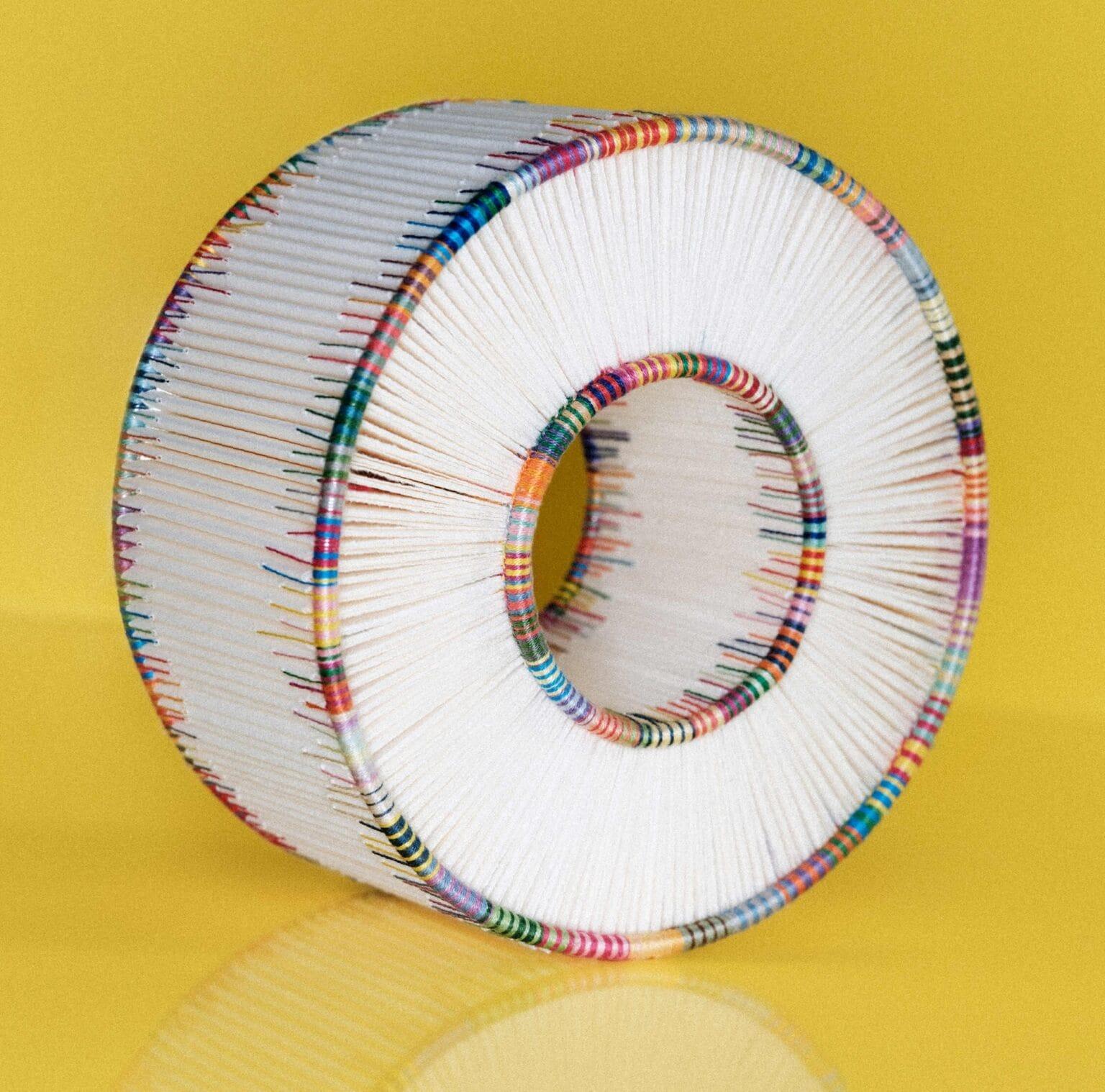 a cylindrical, sculptural form made of folded paper that has been bound along the edges with colorful yarn in the tradition of bookbinding