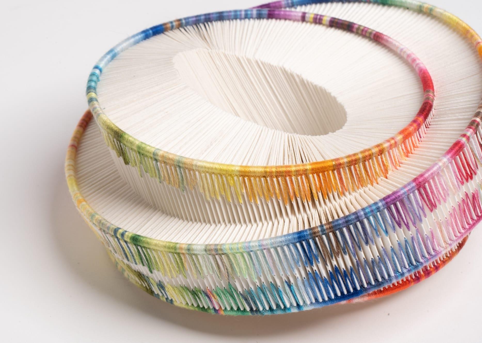 a cylindrical, sculptural form made of folded paper that has been bound along the edges with colorful yarn in the tradition of bookbinding