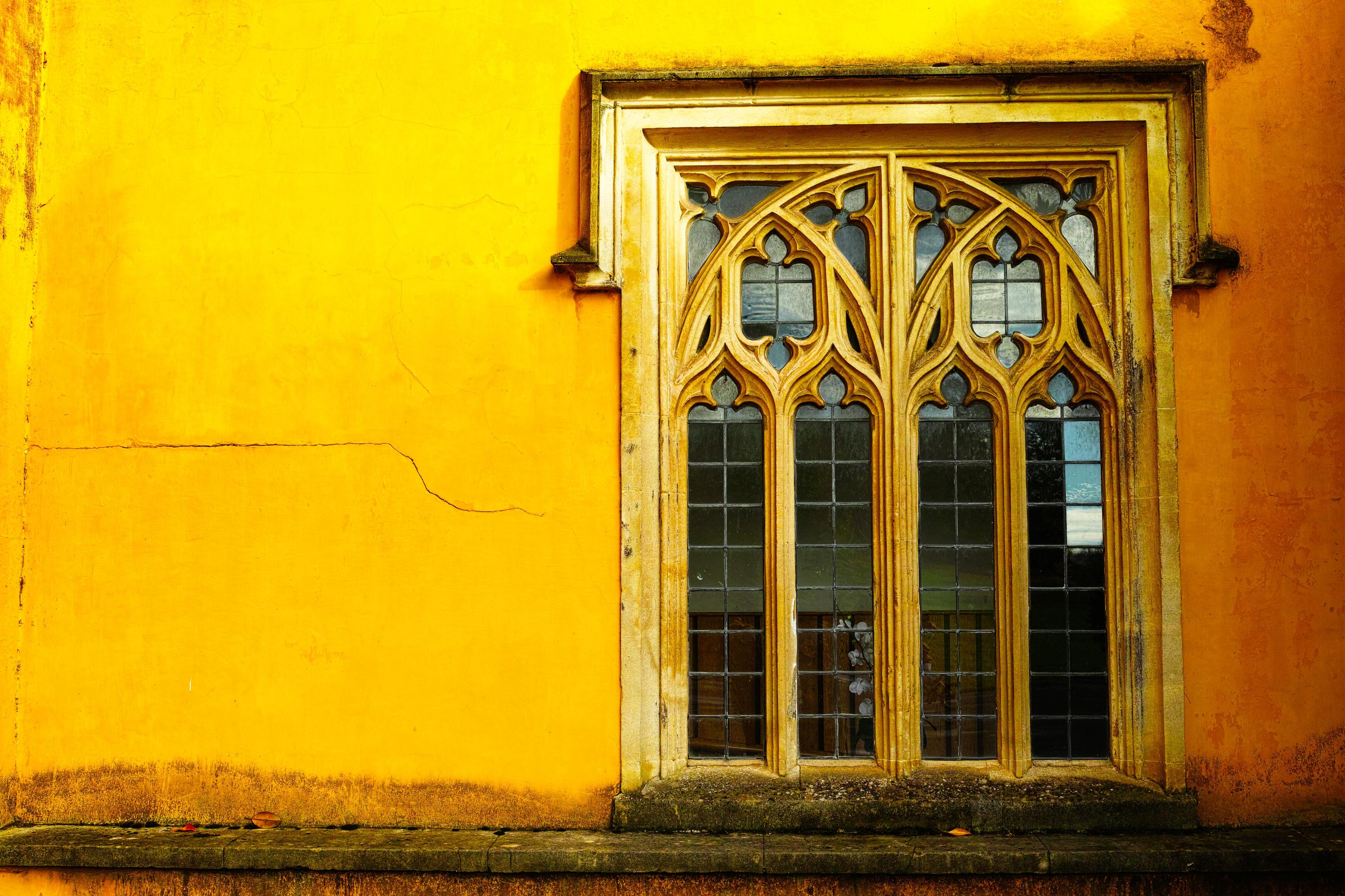 An ornate leaded window with stonework mullions set into a vivid yellow wall lit by sunlight.