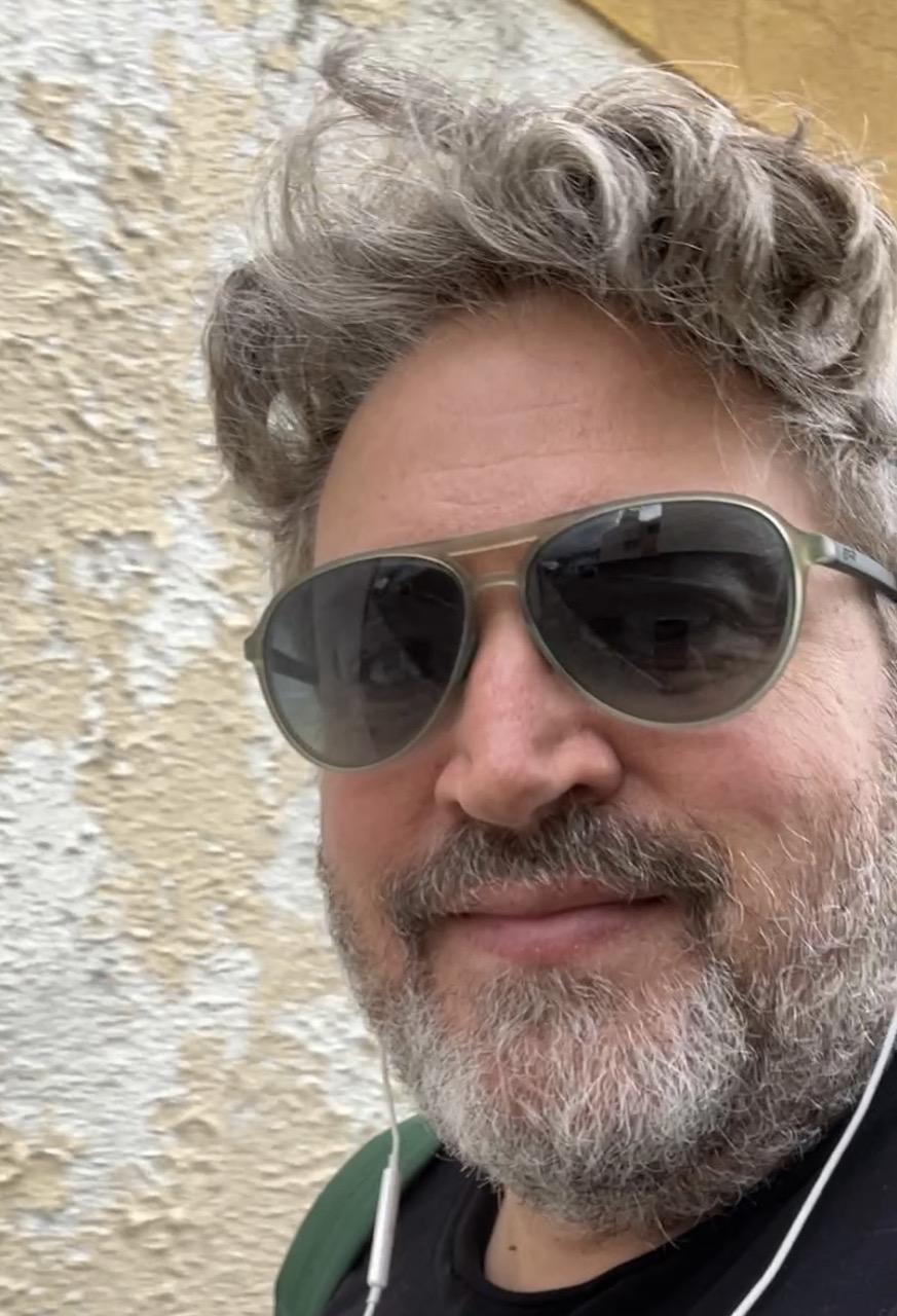 Me, 50ish white man with grey brown hair and grey-brown-white beard looking into camera. Or we presume because I'm wearing dark sunglasses, with light green frames. My hair is a cross between curly and poofy. I'm slightly smiling. I'm wearing white corded headphones and black t-shirt. I'm standing beside a stucco  with flecked paint in white and ecru.