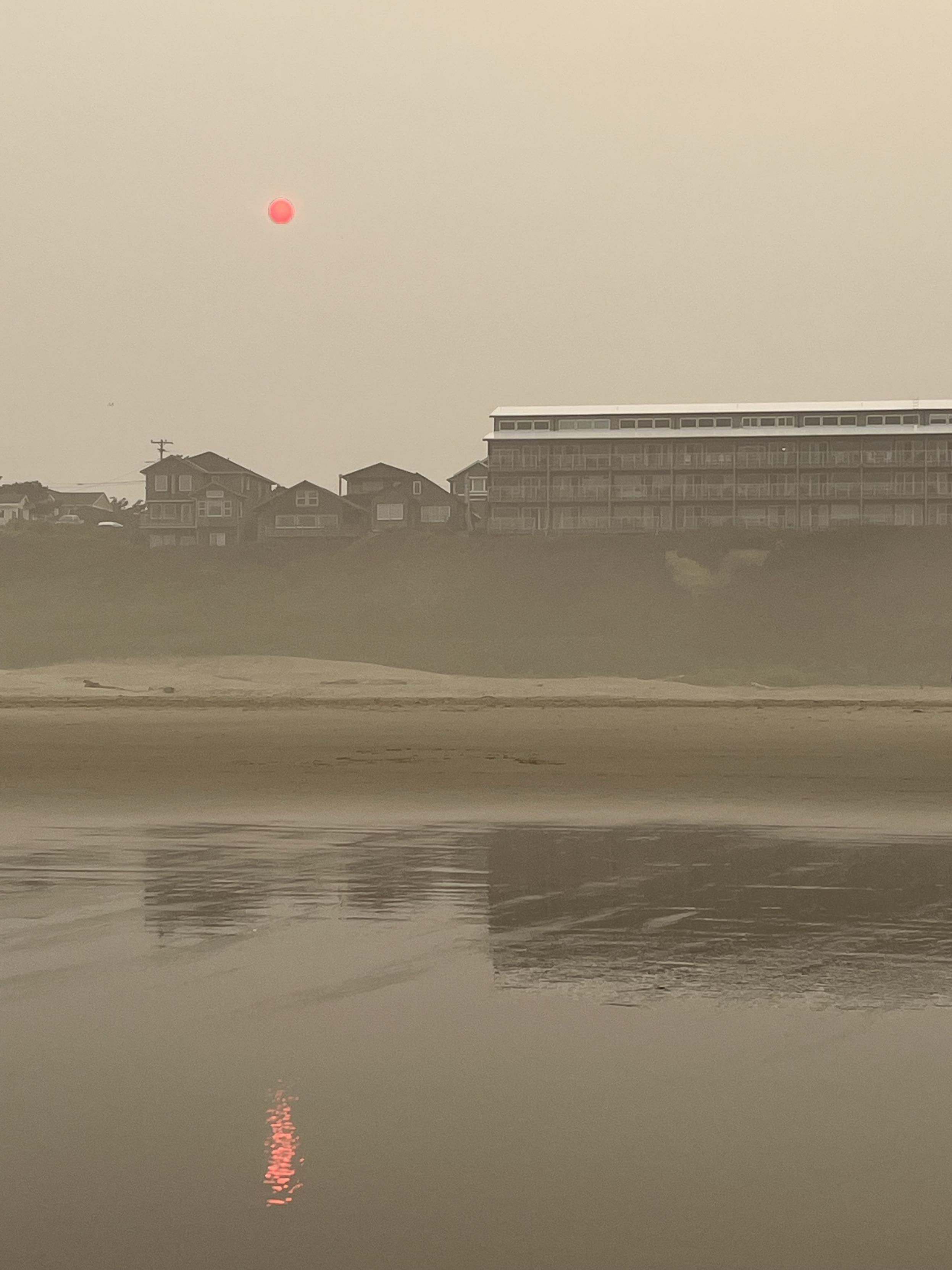 A hazy beach scene features a subdued sun appearing reddish in the sky, casting a reflection on the water. Silhouetted houses and a multi-story building line the shore, with muted colors dominating the landscape due to smoke.