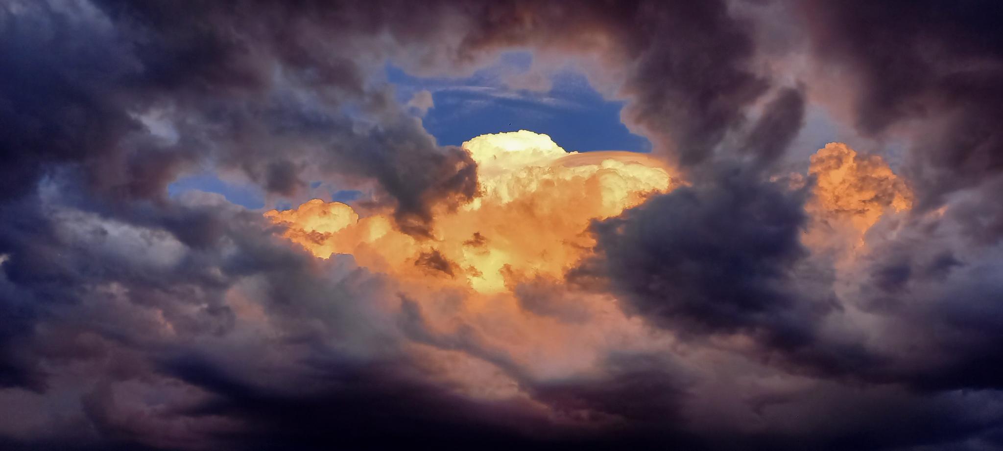 Ciel chargé de nuages très sombres avec une percée par laquelle apparaît un peu de ciel bleu sombre et des gros nuages illuminés par la lumière orange du coucher de soleil Ciel chargé de nuages très sombres avec une percée par laquelle apparaît un peu de ciel bleu sombre et des gros nuages illuminés par la lumière orange du coucher de soleil
