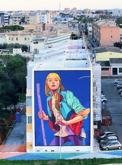 Streetartwall. A beautiful mural of a young woman was sprayed/painted on the outside wall of a three-story building. The painting style is modern, colorful and looks like a good drawing. The background is a strong blue. The young blonde woman is wearing a green, red and blue blouson jacket, a long light-colored T-shirt and purple trousers. She holds a green wooden stick in her hand and looks attentively towards the viewer. One hand rests on the right side of her upper arm, as if someone is trying to stop her.