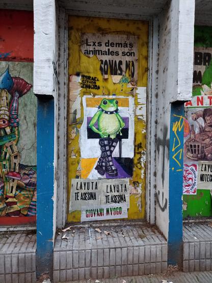Foto de una pared intervenida con distintos carteles superpuestos, rotos, de distintos colores y diseños, y en el centro un dibujo de una rana con pantalones y collar con púas