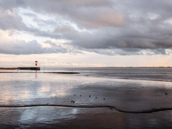 The image shows the Elbe, during low tide. The foreground consists of wet sand and shallow water, reflecting the cloudy sky above. There are small birds scattered across the sand and water.

In the middle ground, there is a narrow strip of water separating the shore from a pier that extends into the sea. At the end of the pier, there is a red and white striped lighthouse.

The background consists of a distant shoreline with wind turbines visible on the horizon. The sky is filled with dark, heavy clouds, with some lighter areas illuminated by sunlight breaking through. The overall atmosphere is calm and serene.