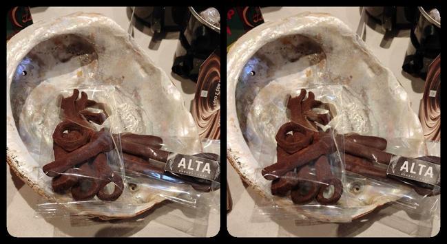 An abalone shell with chocolate packaged in clear cellophane. The chocolate is moulded to look like wrenches, nuts and bolts. It has been dusted with cocoa so it looks rusted. Stereoscopic, parallel view.
