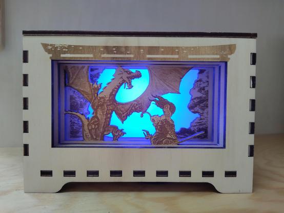 A wooden lightbox showing the with Pepper and its cat on a broom on the foreground, fleeing from an angry dragon ready to attack in the middle layer, with clouds in the background. The scene is backlit in light blue. The box itself has a japanese portal engraved above the scene