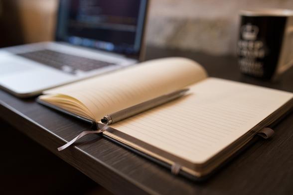 Un carnet ouvert avec un stylo posé dessus. À sa gauche, flouté, un ordinateur portable gris métallisé aux touches noires. À droite, dans le fond, une tasse avec une variante de la mention bien connue « Keep calm and... ».