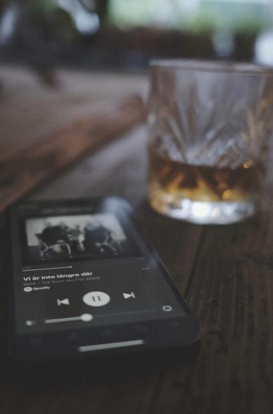 En smartphone på ett bord i trä. På skärmen syns låten ”Vi är inte längre där” av Kent spelas och ur fokus strax bakom telefonen står ett glas whisky.