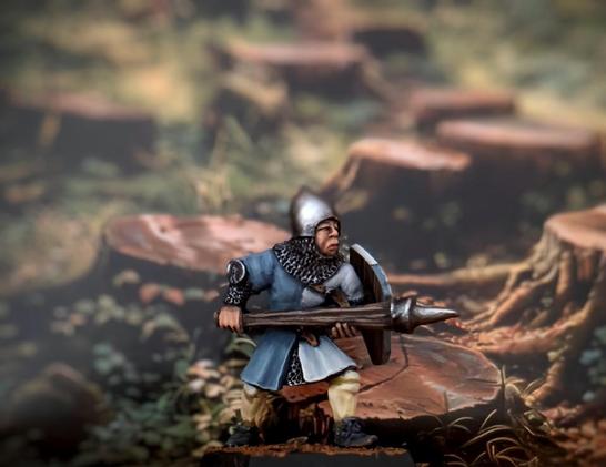 Miniatura de Warhammer de un hombre de armas bretoniano de la tercera edición del juego (sí, 35 años tiene esta miniatura), vestido a lo guerra de los 100 años con una especie de lanza, en realidad un palo con un pincho al final (lo estuve leyendo, era un arma para atacar a los caballos y desmontar a sus jinetes), casco, cota de malla, escudo y la heráldica es gris-azulado y blanco. De fondo hay un bosque talado.