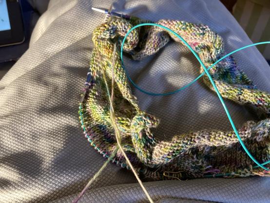 Ein angefangener Loop aus einem gräulichen Garn mit grünen und lila Sprenkeln.