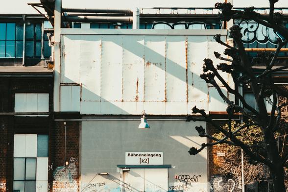 A photo of the stage entrance [k2] from the outside. A large, metal wall hangs above the entrance, with a simple industrial lamp hanging in the center of the frame. Harsh shadows are cast across the wall.