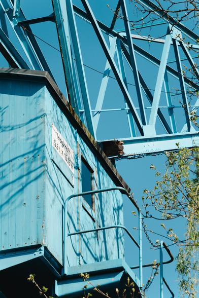 A closeup shot of an old blue crane against a blue sky. The crane cab is built from light blue wooden planks