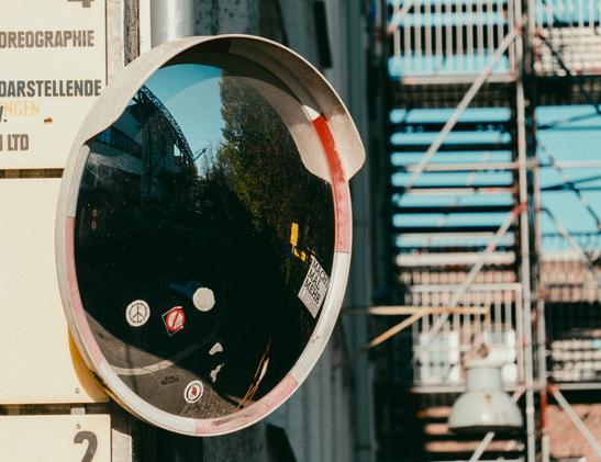 A closeup shot of a road mirror with some stickers on it. industrial buildings are visible both in the reflection in the mirror, as well as in the background around the mirror