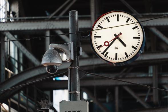 A close shot of a dirty train station clock, next to an old security camera. The picture looks nearly monochrome due to all the metal being the same blue-ish gray. The only bit of color is the red second hand of the clock, as well as a slight red reflection on the rim of the clock.