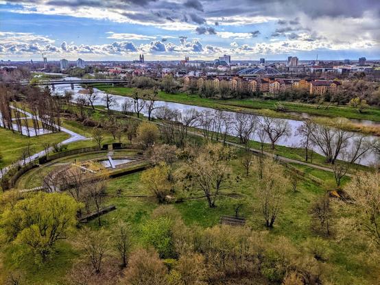 An elevated, wide-angle view shows a cityscape intersected by a winding river. The sky above is a dramatic mix of dark, heavy clouds and patches of bright blue. Below, the river flows gently through a park-like area with bare trees and green spaces. A bridge spans the river in the mid-ground, connecting the foreground greenery to a dense urban area in the distance. This urban landscape features a variety of buildings, including some with distinctive red roofs and taller structures that punctuate the skyline. Footpaths meander through the parkland alongside the river, suggesting a recreational area within the city. The overall mood is one of a city embracing its natural waterways and green spaces, even under a somewhat overcast sky.