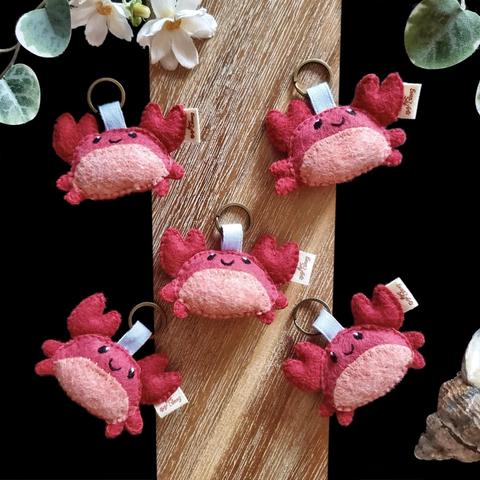 Five little handmade crab keychains photographed on a black and wooden background with a few decorative plants and items.  The keychains are attached to bronze key rings with blue cotton ribbons attached to their heads.  The crabs are red with a pink bellies, two big pincers raised over their heads and two little feet on each sides. Small smiles and little black eyes are embroidered on them.