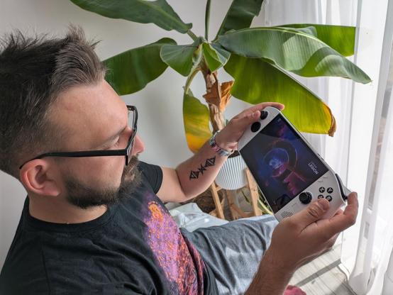 Drugie zdjęcie pokazuje tę samą osobę siedzącą w podobnej pozycji, trzymającą konsolę Lenovo Legion Go. W tle widoczna jest duża roślina doniczkowa z szerokimi liśćmi oraz jasne zasłony. Tatuaż na lewym przedramieniu i zegarek na lewym nadgarstku są dobrze widoczne. Osoba ubrana jest w czarną koszulkę z kolorowym nadrukiem.
