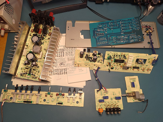 Assorted PCBs from inside a hifi amplifier sitting on an electronics workbench. At the edges of the image, parts of some electronics equipment (power supply, digital multimeter, oscilloscope) can be seen. The topsides of the PCBs are off-white, while the one visible bottom (solder) side is blue.