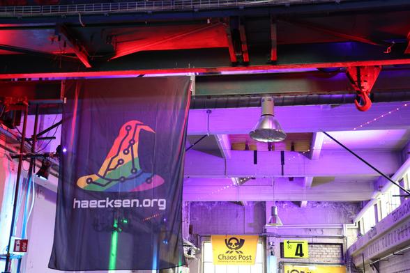 The Heacksen flag, a witch hat in rainbow colors with circuit board traces on it. A Chaos Post sign can be seen in the background.