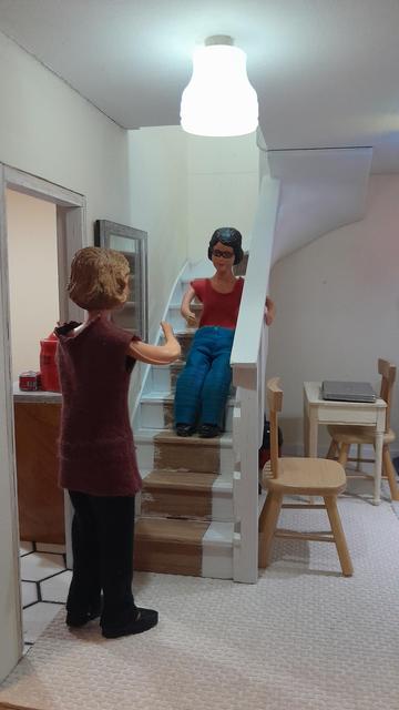 A 1:12 scale diorama of Daisy's living room, looking up at the open plan stairs. The stair treads are painted white at each end but bare wood in the middle where there should be carpet. The kitchen door is on the left at the bottom of the stairs, and there is a wooden chair and a small table to the right of the stairs. The walls are magnolia and the carpet is cream.

Harriet, a Caucasian woman with dark hair and wearing a red tee shirt and blue jeans, is sitting on the stairs holding onto the wooden railings, having been sliding down the stairs on her bum.

Daisy, a Caucasian woman with fair hair and wearing a burgundy top and black trousers, is standing in the kitchen doorway looking up the stairs at Harriet.