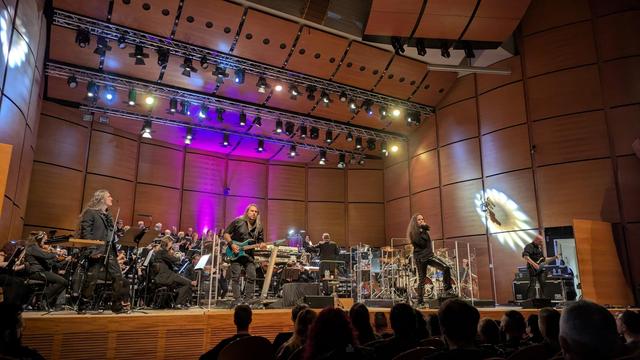 The image depicts a live concert performance in a large auditorium with wooden paneling on the walls. The stage is set with a band performing alongside an orchestra. The band consists of four members: a vocalist on the left, a guitarist in the center, another vocalist on the right, and a bassist at the far right. The vocalist on the left has long, wavy hair and is wearing a black jacket. The guitarist is playing a teal electric guitar and is also dressed in black. The vocalist on the right has long hair and is wearing a black outfit. The orchestra is seated behind the band, with various instruments visible, including brass, woodwinds, and percussion. The stage is illuminated with colorful lights, predominantly purple and white, creating a vibrant atmosphere. The audience is visible in the foreground, with their heads and shoulders shown, indicating their engagement with the performance. The lighting design includes spotlights and colored lights, enhancing the visual appeal of the concert