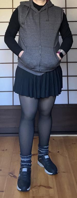 A person wearing black fake transparent tights, a very short plaited skirt, a black shirt and a grey sleeveless vest