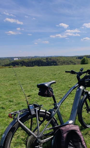 Grüne Wiese mit weitem Ausblick auf entfernt stehende Bäume, blauer Himmel. Im Vordergrund ein abgestelltes Fahrrad, am Gepäckträger klemmt ein Handfunkgerät mit langer Antenne