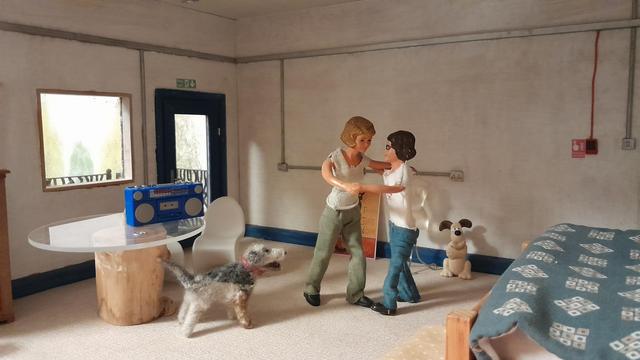 A 1:12 scale diorama of Harriet's bedroom. The walls are grey with electrical conduits. There is a window on the left with a blue glazed fire escape door next to it. The end of the bed is just visible on the right and there is a circular glass table with two white Panton chairs. On the table is a blue portable cassette radio player, an early 80s 'boom box'.

Harriet, a Caucasian woman with dark hair and wearing a white tee shirt and blue jeans, is dancing with Daisy, a Caucasian woman with fair hair and wearing a grey tee shirt and green trousers. 

Monty Dog, Harriet's grey and black dog,  is standing under the table, watching but keeping out of the way.