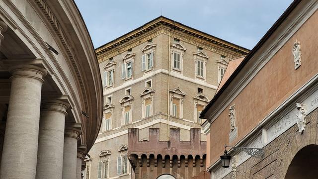 A 9:16 landscape photograph showing, on the left, the external side of Bernini's colonnade for Saint-Peter's square, on the right, the Vatican wall's gate leading north towards piazza del Risorgimento, and in the center, a big corner of the Apostolic palace