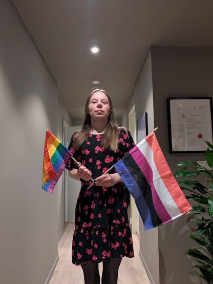 A woman stands in her apartment.

She is holding a LGBTQ+ flag and a genderfluid flag. 

In the foreground, the woman stands in a hallway with an indoor plant visible to the right and a certificate on the wall behind.