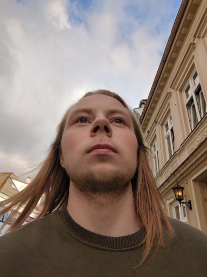 The image features a person standing outdoors. The individual is the central focus, with a low-angle perspective that emphasizes the sky and the surrounding buildings. In the foreground, the person is wearing a dark-colored sweater, while the background reveals a traditional building with windows and an outdoor lamp.