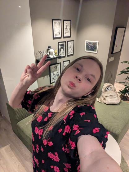 A woman in their mid-twenties, poses for a selfie in her apartment.

Behind them, a shelf displays various figurines and framed artwork.

In the foreground, a sofa and a table can be seen in her living room.