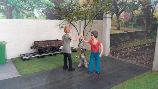 A 1:12 scale diorama, of the courtyard at the front of Harriet's workshop. There is a tarmac drive, with lawn on either side, leading to a pair of tall stone gate posts on the right. A high buff stone wall surrounds the courtyard. There is a small apple tree in the corner, and an old narrow gauge railway wagon on a short length of track is against the wall. Outside the courtyard there is a cobbled track with a grass verge and a low stone wall with mature trees on a green beyond.

Harriet, a Caucasian woman with dark hair, glasses, and wearing a red tee shirt and blue jeans is standing on the drive.

Daisy, a Caucasian woman with fair hair, and wearing a green tee shirt and black trousers, is also standing on the drive.

The two women are almost dancing around each other, high fiving with their right hands.

Monty Dog, Harriet's grey and black dog is standing on his hind legs, between them, trying but unable to reach to join in the high fiving.