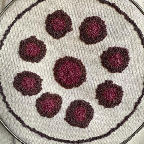 Nine circles of pink and purple French knot flowers, 8 in a circle surrounding a larger and more pink bloom in the middle. They're on naturally colored cotton artist's canvas and encircled by a single circle of darker purple French knots. It's still in its aluminum spring tensioned embroidery hoop.