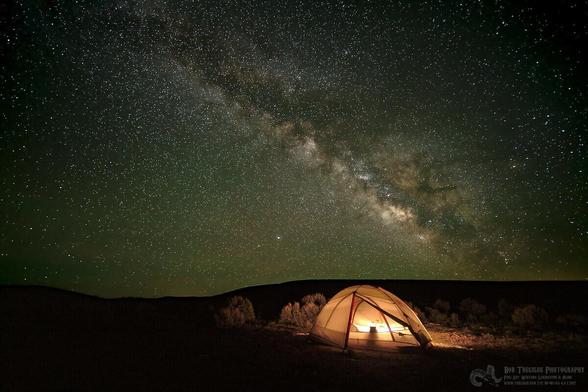 A photo of a brightly lit tent, a cat sitting inside looking out wondering why his human is messing around outside when it is so cold, and the Milky Way above the tent in the background.