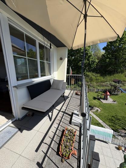 A balcony scene featuring a sunshade, a cushioned bench, and a planter with green plants. In the background, a well-maintained garden can be seen, along with a small red structure, grass, and some decorative elements. Bright sunlight illumin