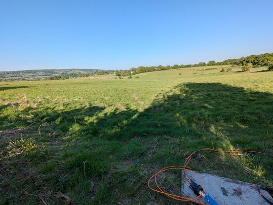 A photo of several large fields dropping slightly revealing a hill in the distance. Blue sky and green grass is abundant.
Bottom right of the shot is a chamber lid with some fibre equipment on the top. Orange tubes come from the equipment.