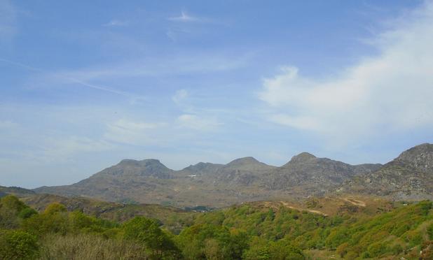landscape photograph by Remy Dean of distant hazy mountains beyond a series of valleys where the trees and other foliage has a bright fresh green abundance - the sky is pale blue with wispy clouds